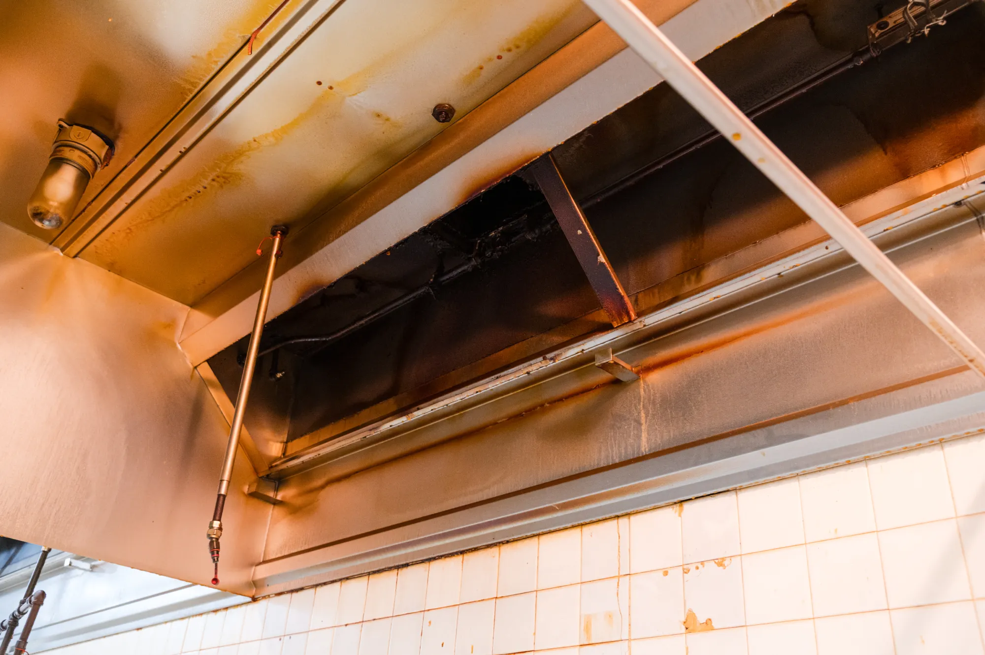 Dirty commercial kitchen exhaust hood with grease stains and buildup on metal surfaces above tiled wall.