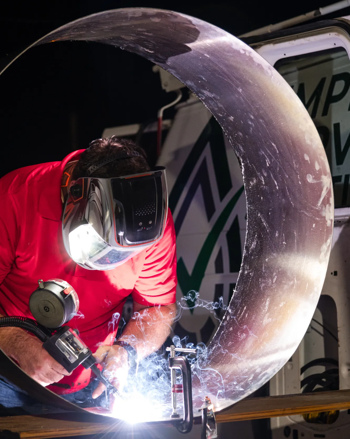 A person welding a large metal pipe, wearing a protective mask and red shirt, with sparks and smoke visible.
