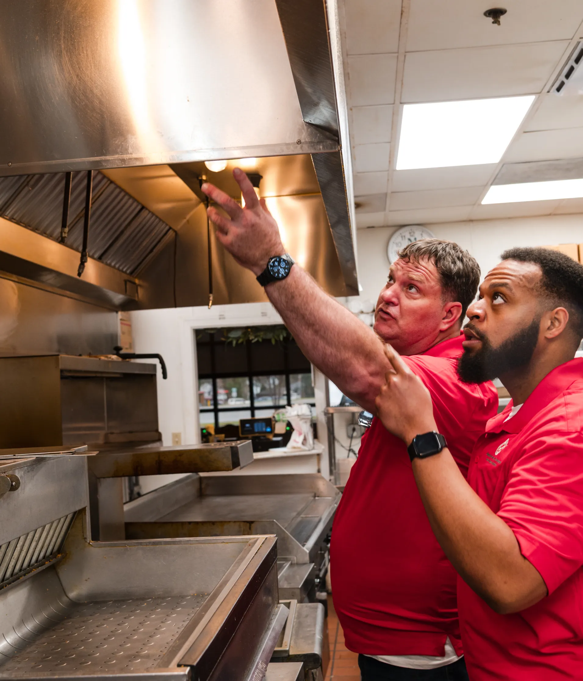 home-1 Two men in red shirts inspect a kitchen hood in a commercial restaurant kitchen.