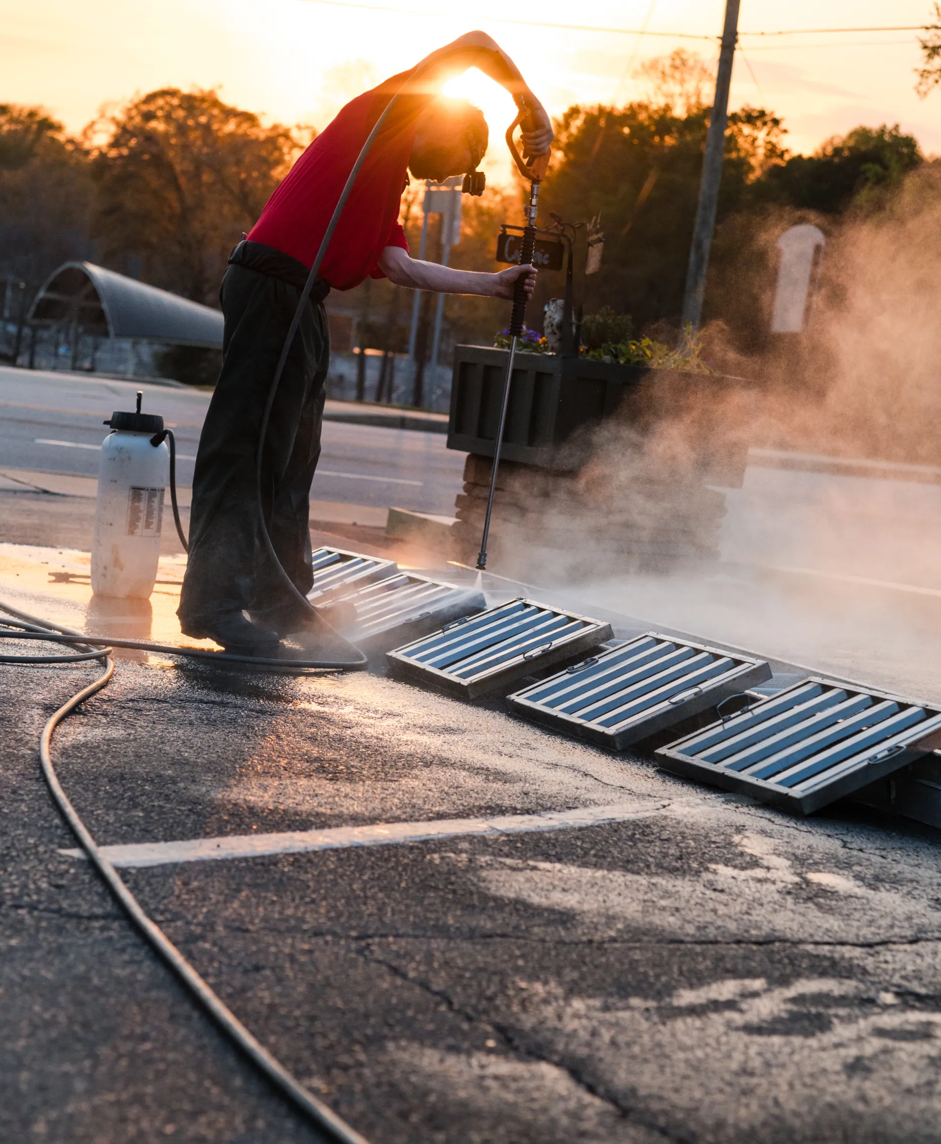 hood-4 A person power washing metal grates outside at sunset, with mist rising and hoses on the ground.