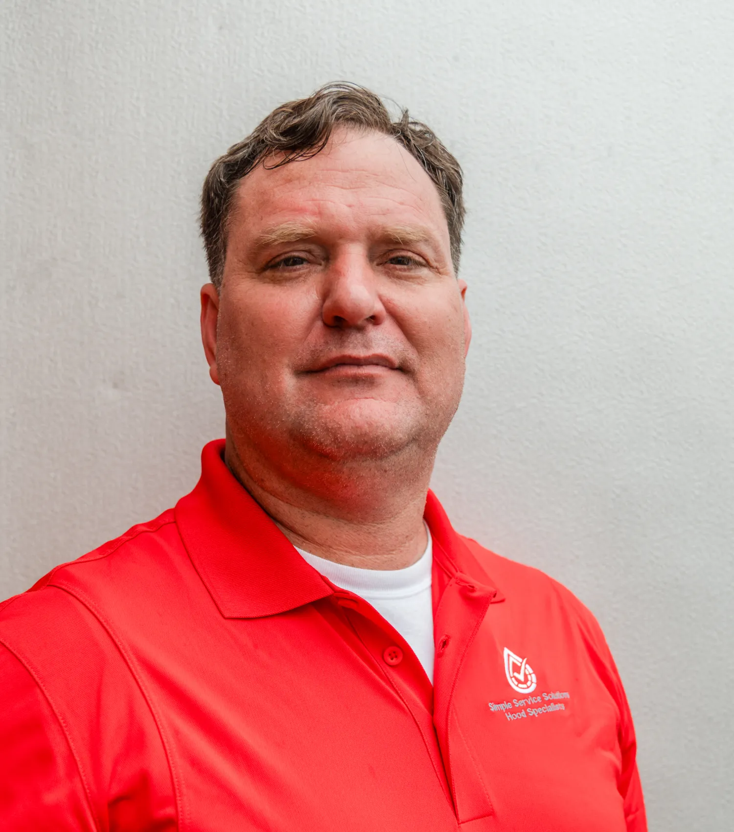 team-1 Man in a red polo shirt standing against a plain white wall, looking at the camera with a neutral expression.