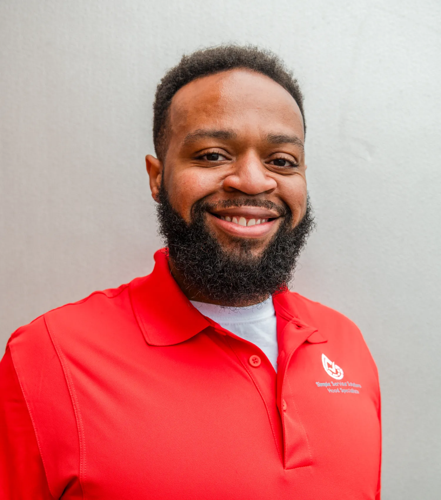 team-2 Smiling man with a beard wearing a red collared shirt, standing in front of a light gray background.