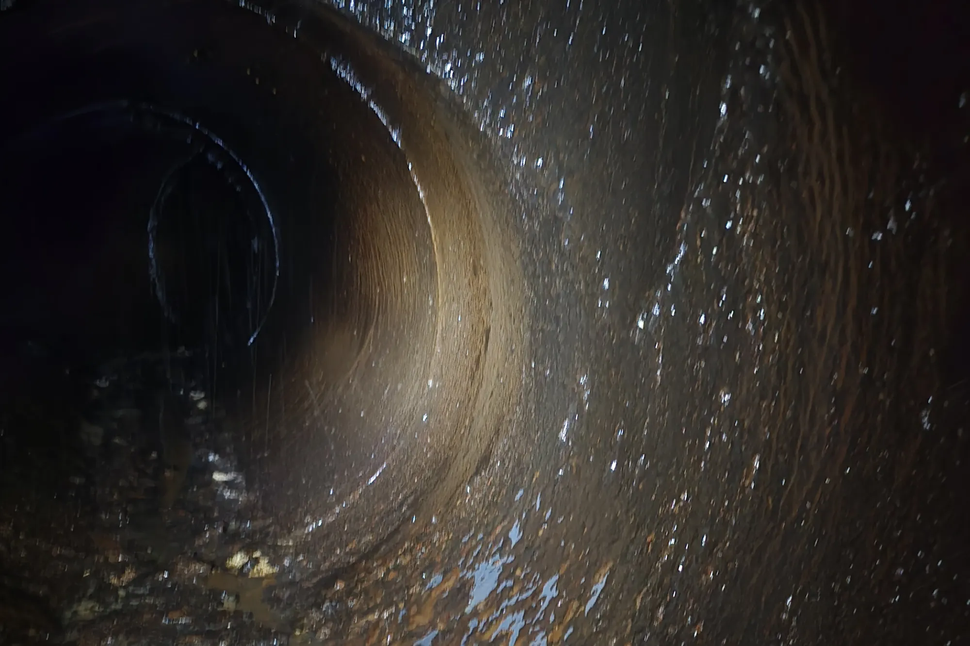 A dark, wet, circular tunnel with water and debris along its rough, grimy surface.